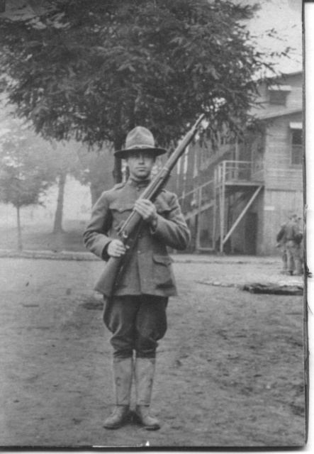 photo: WWI photo of Norman in military uniform and hat hold a riffle. Appears to be in front of some barracks.